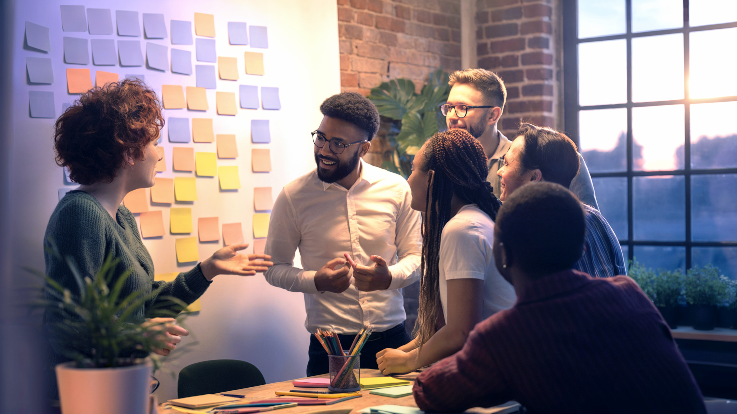 Eine Gruppe von Menschen steht vor einer Wand voller bunter Notizzettel und spricht lebhaft miteinander. Das warme Licht fällt durch große Fenster und symbolisiert gemeinsame Gestaltung und neue Wege.